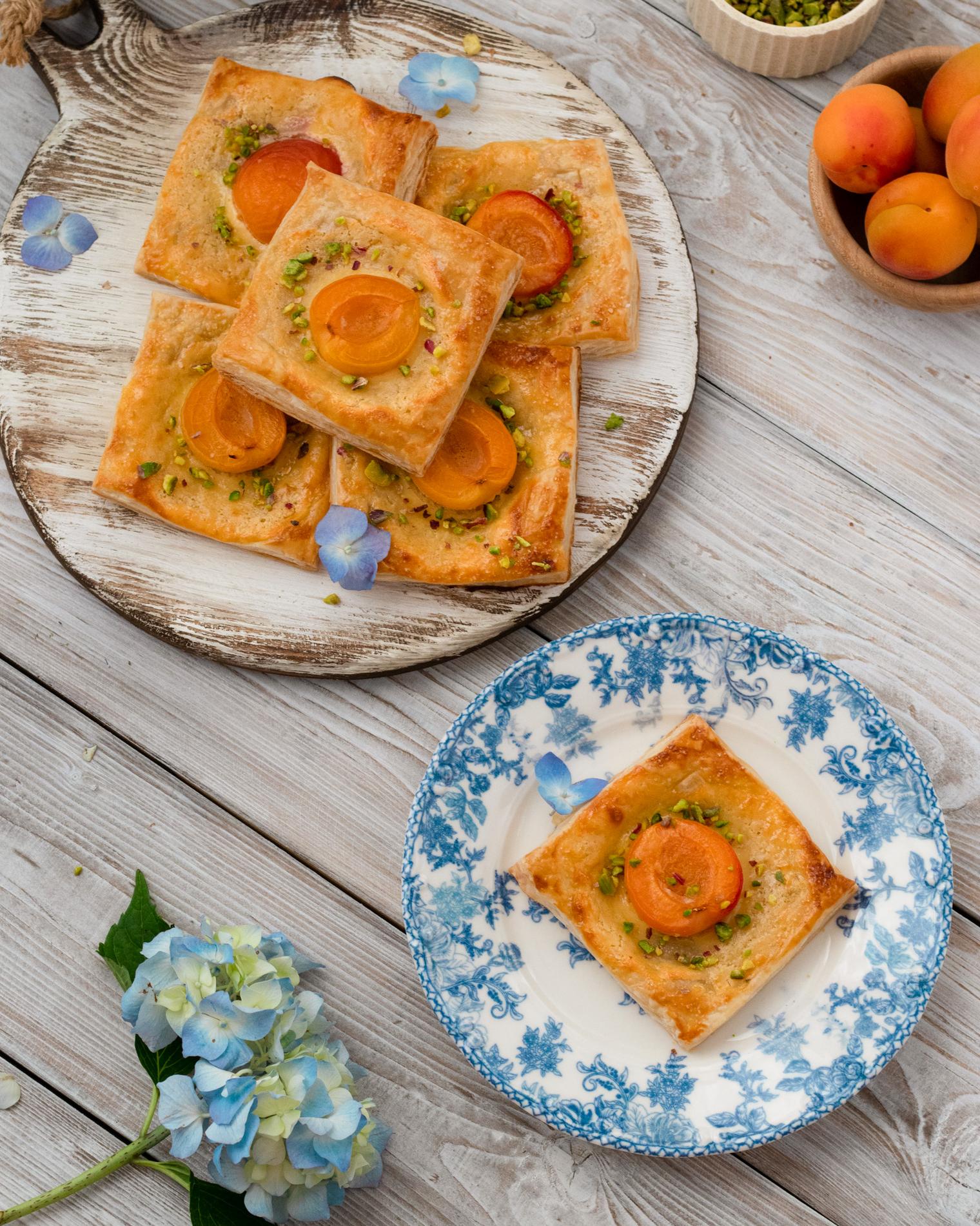 Top-down view of golden square puff pastries each topped with a halved glazed apricot and scattered crushed pistachios, arranged on a distressed white round board and one on a blue-and-white floral plate; soft pastel styling with scattered blue hydrangea petals, a small wooden bowl of whole apricots, and a whitewashed plank table under warm natural light, emphasizing flaky texture, warm orange tones and cool blue accents.