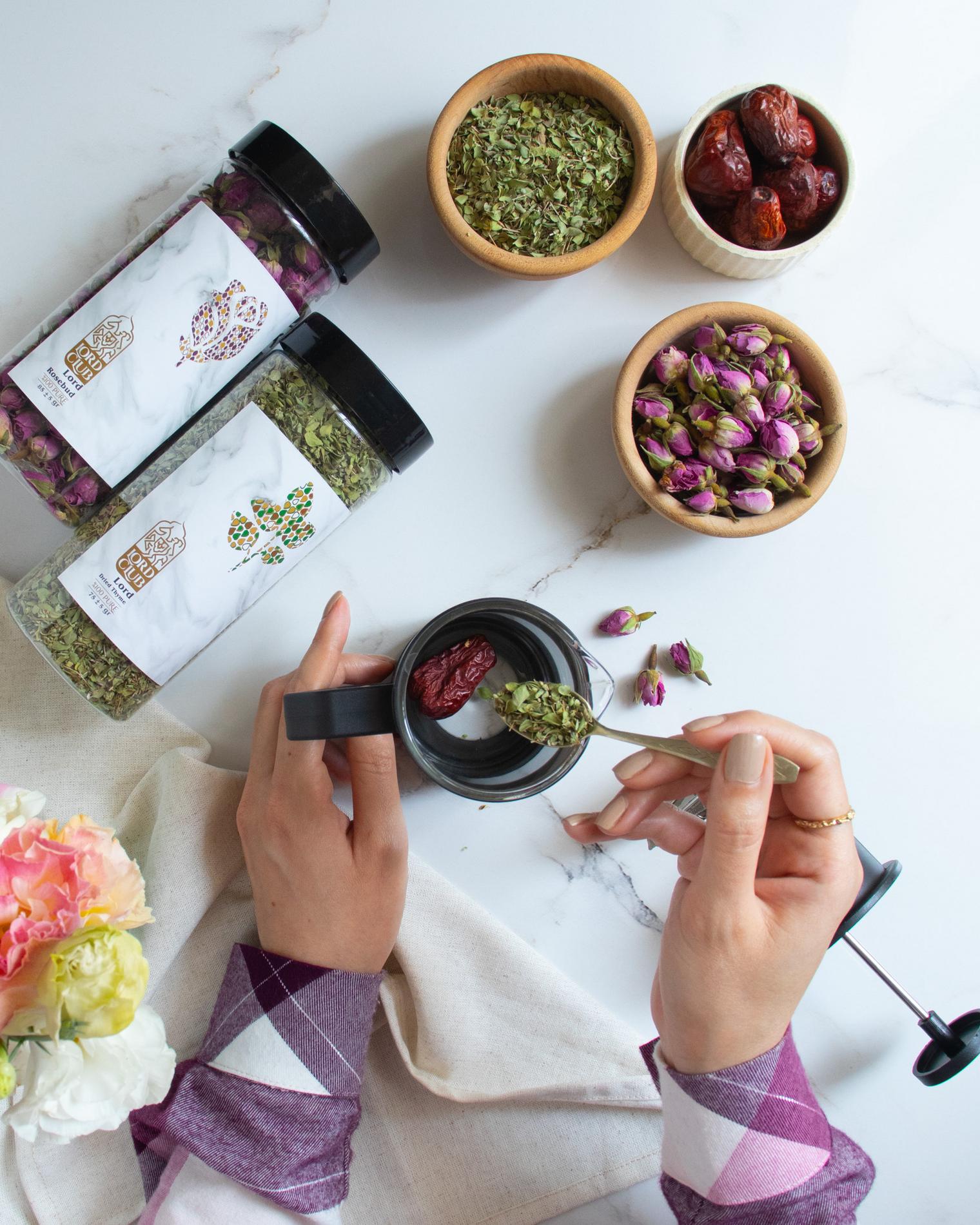 Top-down flatlay of a botanical tea ritual on a white marble surface: two hands with neutral manicure and a thin gold ring hold a black-handled mug and spoon a heap of dried green herbs into it; a red dried date sits inside the mug. To the left are two clear jars with black lids and minimalist marble labels filled with dried herbs and rosebuds. Three small wooden bowls at the top contain loose oregano-like herbs, whole dried dates, and a pile of pink rosebuds. A cream linen napkin, soft pastel flowers at the bottom-left, and a French-press plunger at the right edge complete the clean, natural composition.