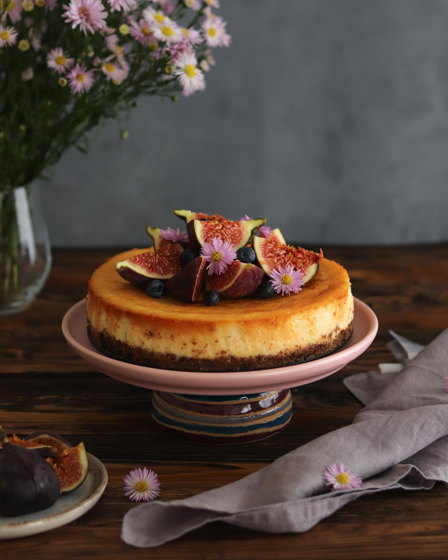 Caramel-topped cheesecake on a pink ceramic stand, decorated with halved fresh figs, blueberries and small pink daisy-like flowers; set on a dark wooden table with a folded mauve linen napkin to the right, a small plate of extra figs at lower left and a blurred vase of pink daisies in the upper left against a textured gray backdrop. Shot at eye level with soft natural window light from the left and a shallow depth of field that highlights the cake's creamy texture, fig interiors and delicate floral accents.