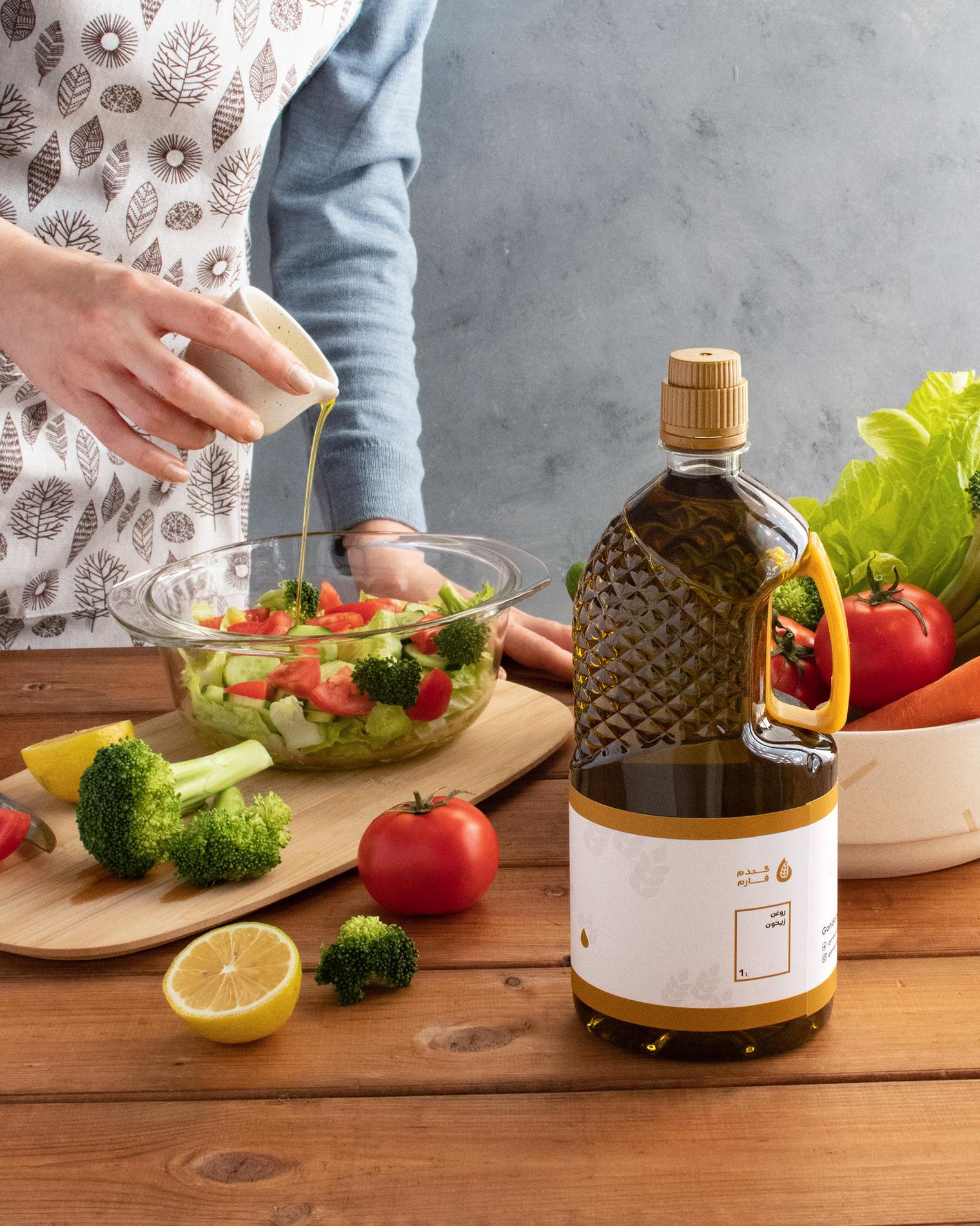 Close-up editorial still life of a hand pouring a stream of golden olive oil from a small white pitcher into a glass bowl of fresh salad. The salad contains torn lettuce, halved cherry tomatoes and broccoli florets; a halved lemon, whole tomatoes and broccoli pieces sit on a warm wooden table. A large textured olive oil bottle with a yellow handle and a blank white label is placed prominently in the foreground to the right. The person pouring wears a blue long-sleeve top and a white apron with a delicate leaf pattern; the scene is lit with soft natural light against a muted grey backdrop, creating a warm, rustic and minimal aesthetic.